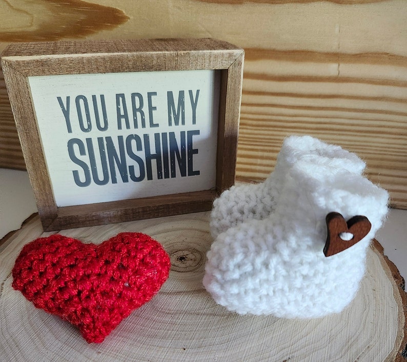 Op de afbeelding: Een witte gehaakte babyschoen met een houten hartknop, een rood gehaakt hart en een ingelijst bord met de tekst "YOU ARE MY SUNSHINE". De items worden gepresenteerd op een houten oppervlak.