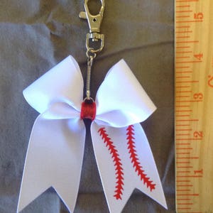 May include: White cheer bow with red baseball stitching, attached to a silver key chain.