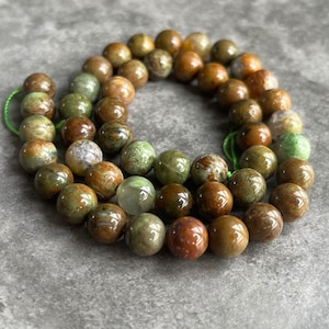 Puede incluir: Hilo de cuentas redondas de piedras preciosas en tonos marrón, verde y crema. Las cuentas están ensartadas en un cordón verde, para un collar o pulsera. Las cuentas tienen un acabado pulido y brillante.