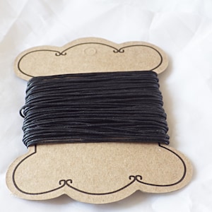 May include: A spool of black waxed cotton cord. The cord is tightly wound on a decorative, cloud-shaped cardboard spool. The spool has decorative swirls at the bottom. The background is a white, textured fabric.