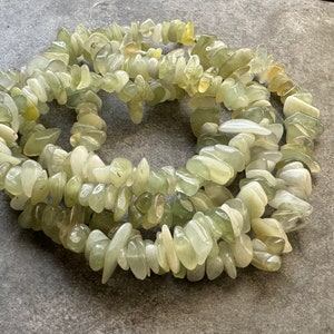 Pu&ograve; includere: Una collana di perle a scaglie levigate, di forma irregolare e nei toni del verde pallido, bianco traslucido e giallo tenue. Le pietre lucidate sono infilate in una disposizione circolare, con una consistenza liscia, lucida e forme organiche.