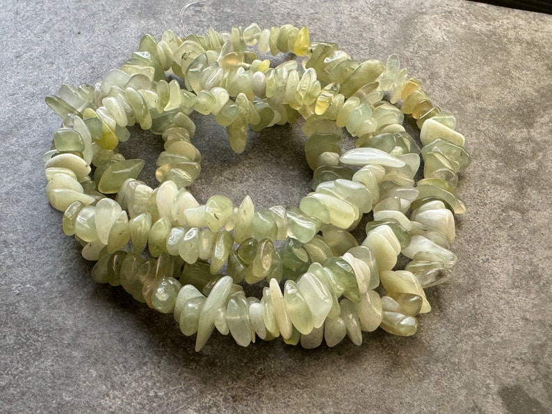 Pu&ograve; includere: Una collana di perle a scaglie levigate, di forma irregolare e nei toni del verde pallido, bianco traslucido e giallo tenue. Le pietre lucidate sono infilate in una disposizione circolare, con una consistenza liscia, lucida e forme organiche.
