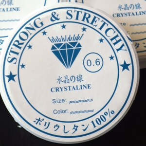 Könnte beinhalten: Ein weißes rundes Etikett mit blauem Text und einem blauen Diamantenlogo. Das Etikett lautet "Strong & Stretchy" und "Crystaline". Das Etikett enthält auch den Text "Größe:" und "Farbe:" mit wellenförmigen Linien darunter. Das Etikett enthält auch den Text "0.6" und "Polyurethan 100 %".