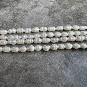 May include: Four strands of white, irregularly shaped pearls are shown against a grey background. The pearls have a lustrous sheen and vary in size and shape, with some appearing more elongated and others rounder.