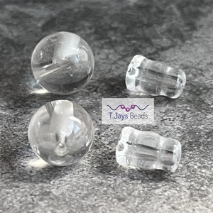 May include: Four clear glass beads, two round and two cylindrical, on a grey textured surface.  T.Jays Beads is printed on a white label.