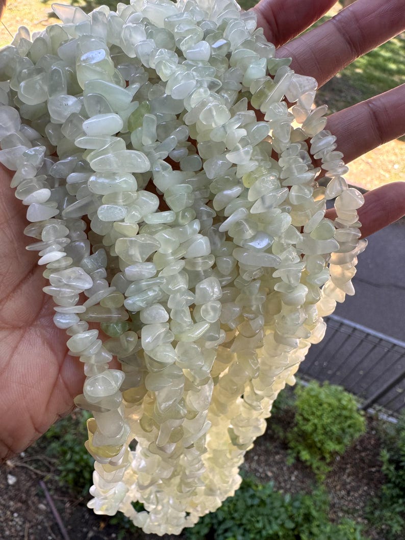 Pu&ograve; includere: Una collezione di molteplici fili di scaglie di pietre preziose levigate, di colore verde pallido e traslucide. Le pietre sono di forma e dimensioni irregolari, con una finitura liscia e lucida che cattura la luce, creando un aspetto morbido.