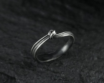 May include: A silver-toned metal ring featuring a thin, multi-layered band design that converges into a small, central knot detail at the top. The ring has a polished finish with subtle dark oxidation in the recessed areas of the band.
