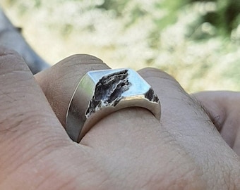 May include: A silver-toned ring with a smooth, polished band and a textured, rugged top surface that resembles a mountain peak. The ring is worn on a finger, and the text "DESIGN 1" is printed in the bottom left corner of the image.