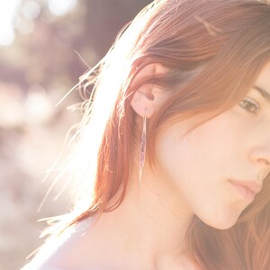 May include: A woman with long, auburn hair wearing a pair of long, slender earrings. The earrings have a linear design with a subtle, light-colored pattern. The background is blurred with sunlight.