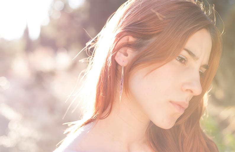 May include: A woman with long, auburn hair wearing a pair of long, slender earrings. The earrings have a linear design with a subtle, light-colored pattern. The background is blurred with sunlight.