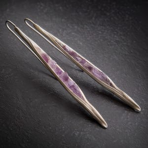 May include: A pair of silver-toned earrings with a long, slender design. Each earring features a central inlay of rough-cut, light purple amethyst stones. The earrings are set against a dark, textured background.