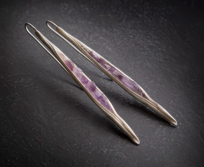 May include: A pair of silver-toned earrings with a long, slender design. Each earring features a central inlay of rough-cut, light purple amethyst stones. The earrings are set against a dark, textured background.