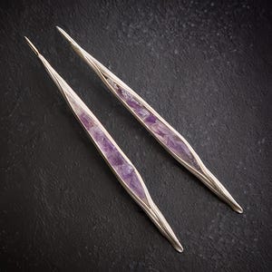 May include: A pair of silver-toned earrings with a long, slender design. Each earring features a central inlay of vibrant purple amethyst stones. The earrings are set against a dark, textured background, highlighting their elegant form.