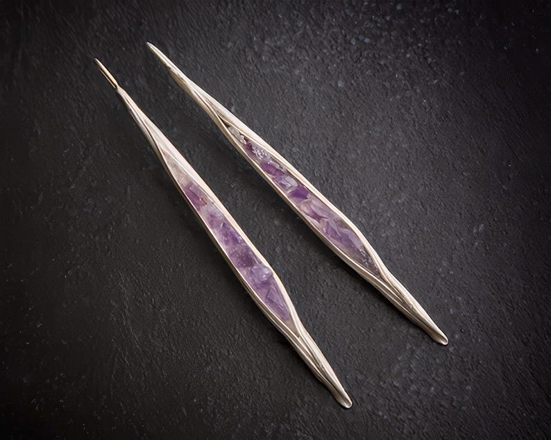 May include: A pair of silver-toned earrings with a long, slender design. Each earring features a central inlay of vibrant purple amethyst stones. The earrings are set against a dark, textured background, highlighting their elegant form.