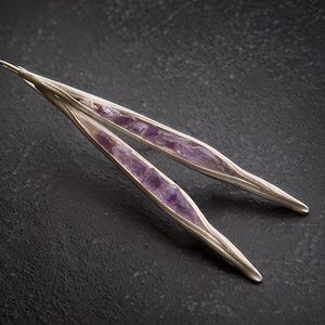 May include: A pair of silver-toned earrings with a long, slender design. Each earring features a central inlay of vibrant purple amethyst stones. The earrings are set against a dark, textured background, highlighting their elegant form.