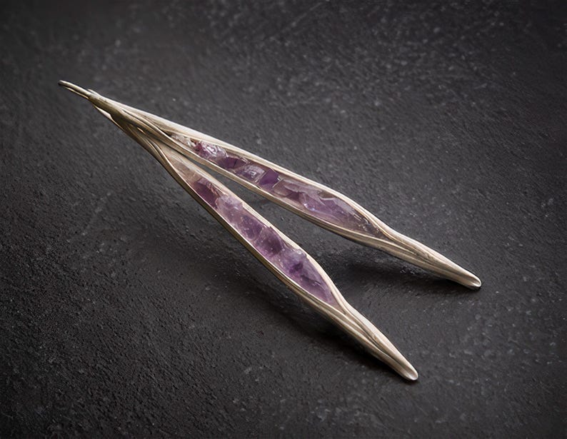 May include: A pair of silver-toned earrings with a long, slender design. Each earring features a central inlay of vibrant purple amethyst stones. The earrings are set against a dark, textured background, highlighting their elegant form.