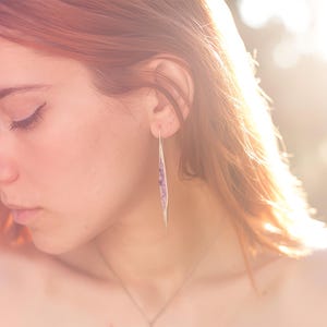 May include: A side profile of a person with red hair, wearing a silver earring. The earring is a long, slender, leaf-like shape with a purple design. The person is looking down, with a nose ring and a necklace.