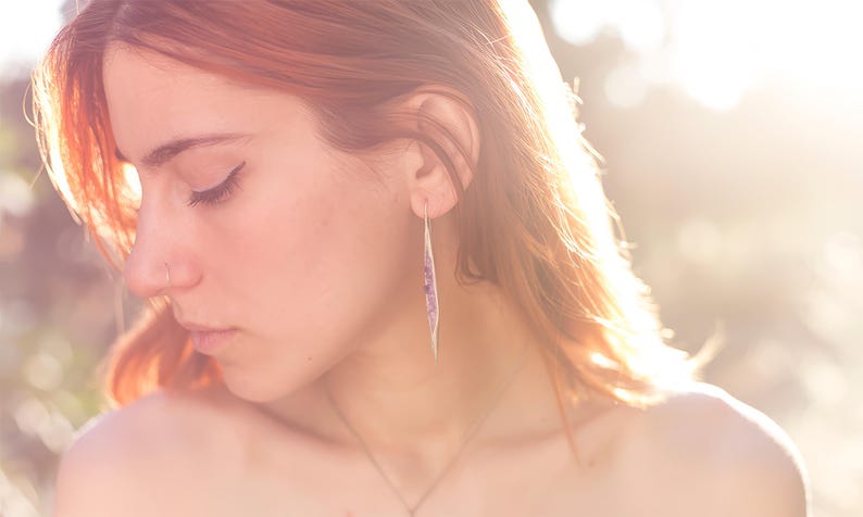 May include: A side profile of a person with red hair, wearing a silver earring. The earring is a long, slender, leaf-like shape with a purple design. The person is looking down, with a nose ring and a necklace.