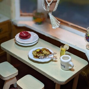 May include: A miniature dollhouse scene with a table set for breakfast. There are two white stools, a white plate with waffles and syrup, a glass of lemonade, and a white mug with the word "Berly" on it. There are also three white plates stacked on top of a red plate.