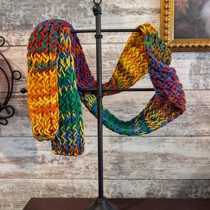 May include: A colorful, hand-knitted infinity scarf displayed on a black metal stand. The scarf features a vibrant mix of red, orange, yellow, green, and blue yarns. The textured knit creates a visually appealing pattern, perfect for adding a pop of color to any outfit.