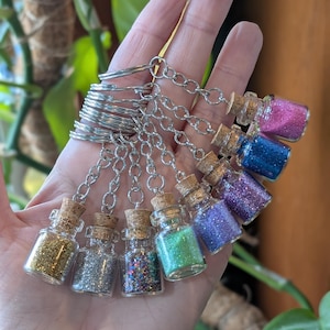 Könnte beinhalten: Ein Schlüsselanhänger mit acht kleinen Glasflaschen, die mit verschiedenen Farben Glitter gefüllt sind. Die Flaschen haben Korkstopfen und sind an einer silbernen Kette befestigt.