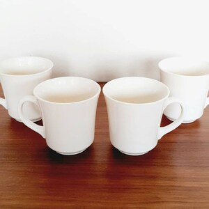 May include: Four white ceramic mugs with rounded handles. The mugs are arranged in a semi-circle on a wooden surface.