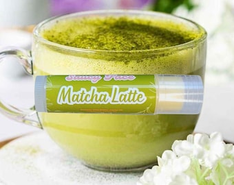Könnte beinhalten: Ein klares Glas mit Matcha Latte, bedeckt mit grünem Matcha-Pulver. Eine Lippenbalsam-Tube mit dem Text "Saucy Face Matcha Latte" befindet sich vor dem Becher. Weiße Blumen sind im Vordergrund.