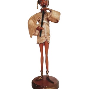 May include: A wooden figurine of a person playing a flute. The figurine is wearing a gold-colored robe and has brown skin. The figurine is standing on a round wooden base.