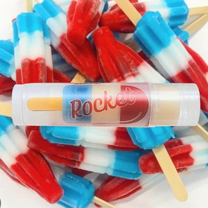 Op de afbeelding: Een doorzichtige tube lippenbalsem met een rood-wit-blauw etiket waarop "Rocket" staat. De tube is omringd door rood-wit-blauwe ijslolly's op houten stokjes.