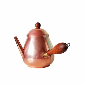 Puede incluir: Una tetera de cobre con asa y pomo de madera. La tetera tiene una superficie brillante y reflectante y un pico para verter. El asa es curva y está unida al lateral de la olla. La tapa tiene un pequeño pomo de madera redondo.