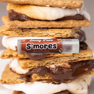 Könnte beinhalten: Ein Stapel S'mores mit Graham Crackern, Schokolade und Marshmallow. Eine Lippenbalsam-Tube mit dem Text "Saucy Face S'mores" liegt oben auf den S'mores. Die Lippenbalsam-Tube ist braun und weiß.