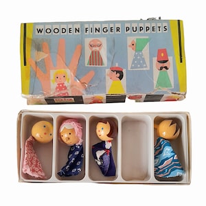 May include: A set of four wooden finger puppets in a white box. The puppets are dressed in colorful fabric and have painted faces. The box has the words "Wooden Finger Puppets" printed on the top.