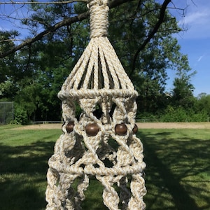 Puede incluir: Un macetero de macramé con un diseño de cuerda blanca y beige. El macetero tiene cuentas de madera y una parte superior decorativa.