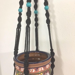 Op de afbeelding: Zwarte macramé plantenhanger met turquoise kralen, die een terracotta pot met een kleurrijk bloemenmotief vasthoudt.