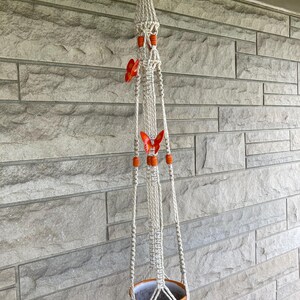 May include: A macramé plant hanger with a decorative ceramic pot. The off-white hanger features orange beads and two orange butterfly accents. The ceramic pot has a colourful, patterned design.