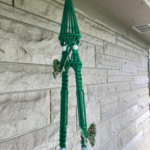 Op de afbeelding: Een handgemaakte groene macramé plantenhouder met witte kralen en vlinderaccenten. De plantenhouder is opgehangen aan een metalen ring en is ontworpen om een ​​bloempot te bevatten. De vlinders hebben een groen en oranje patroon.