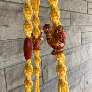 May include: Yellow macrame plant hanger with brown ceramic rooster accents. The hanger features a knotted design and a wooden bead. The roosters are glazed in shades of brown and yellow. A decorative piece for indoor or outdoor use.