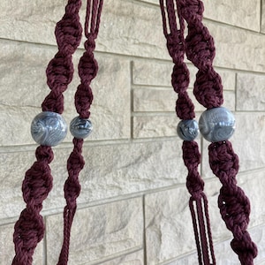 Peut inclure: Un macramé de couleur bordeaux avec des nœuds décoratifs et des perles grises marbrées. Le support est conçu pour contenir un pot, avec un bord turquoise et marron visible en bas. Le fond est un mur de briques de couleur claire.