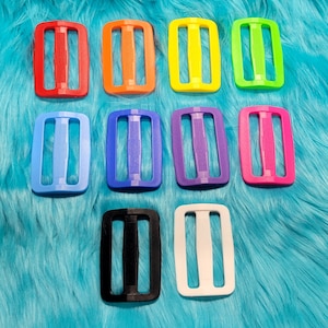 May include: Ten plastic slider buckles in various colors, including red, orange, yellow, green, blue, purple, pink, black, and white. The buckles are all the same size and shape, with two holes on each side.