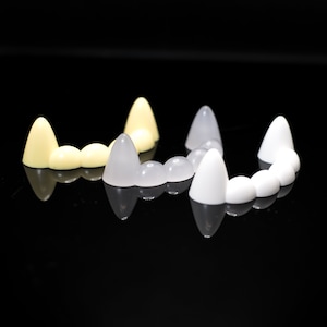 May include: Three sets of plastic vampire teeth, one set is white, one set is off-white, and one set is a darker shade of white. The teeth are arranged in a semi-circle shape.
