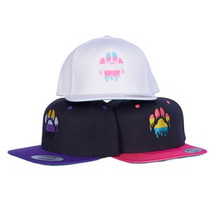Op de afbeelding: Drie baseballpetten met verschillende gekleurde poot afdrukken. De bovenste pet is wit met een roze, blauwe en witte poot afdruk. De middelste pet is zwart met een paarse, gele en witte poot afdruk. De onderste pet is rood met een gele, blauwe en roze poot afdruk.