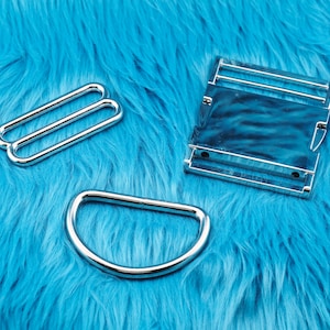 May include: Three shiny silver metal hardware pieces on a bright blue, textured background. The pieces include two rectangular sliders, a D-ring, and a rectangular buckle. These are likely used for bag making or other crafting projects.