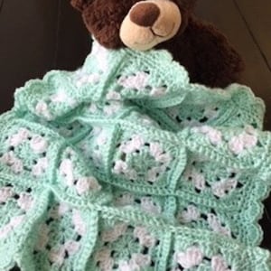 May include: A crocheted baby blanket in a light green and white color scheme. The blanket is made of squares with a delicate pattern and has a ruffled edge.