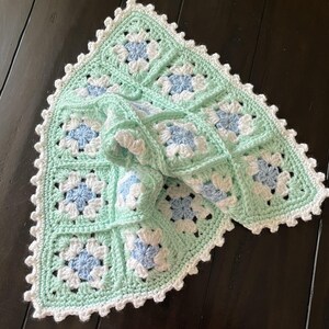 May include: A square crochet blanket made of joined granny squares in light mint green, white, and pale blue yarn. The blanket features a decorative scalloped white border along the edges, with a repeating floral-inspired pattern in each square.