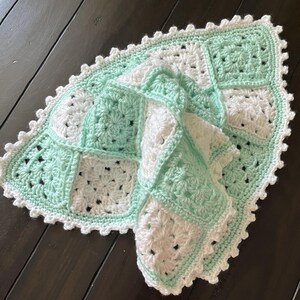 May include: A handmade crochet blanket featuring a repeating pattern of alternating mint green and white granny squares. The blanket is finished with a decorative scalloped edge border in white yarn, creating a textured and soft appearance.