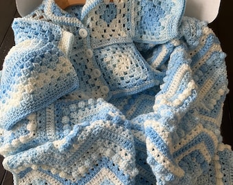 May include: A light blue and white crocheted cardigan and matching blanket set. The cardigan features a granny square pattern with white buttons down the front. The blanket displays a textured chevron and bobble stitch design.