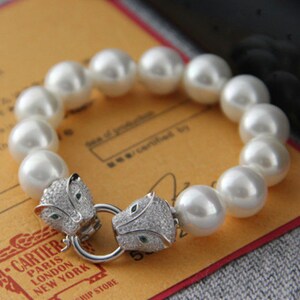May include: A white pearl bracelet with a silver clasp featuring two panther heads with green gemstone eyes.