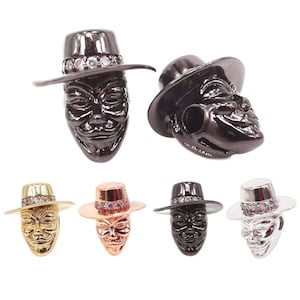May include: Five charm pendants featuring a stylised face wearing a hat with a band of rhinestones. The charms are available in black, gold, rose gold, and silver.