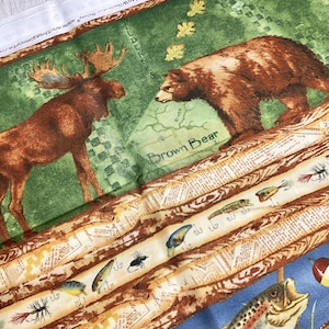 May include: A fabric panel with a green background featuring a brown bear and a moose. The panel also includes a brown and white striped pattern with fishing lures and the text "Brown Bear".