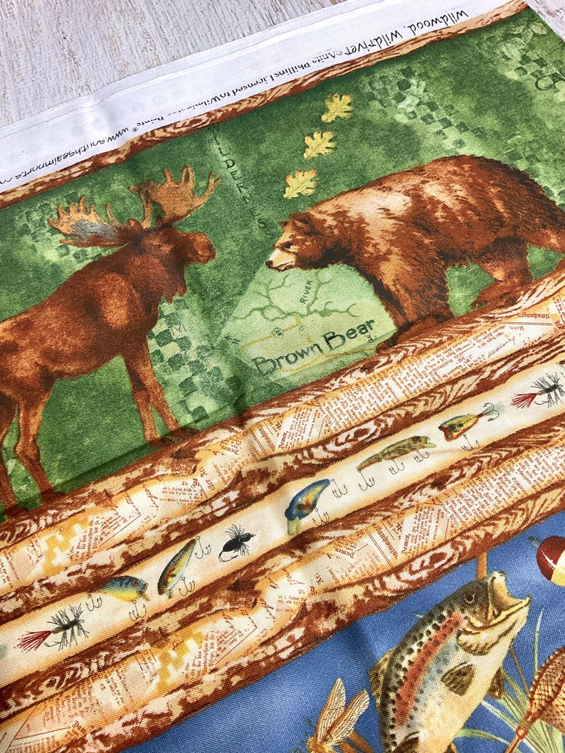 May include: A fabric panel with a green background featuring a brown bear and a moose. The panel also includes a brown and white striped pattern with fishing lures and the text "Brown Bear".